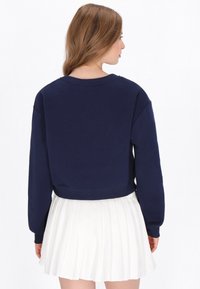 Sötétkék, rövidített pulóver hosszú ujjakkal, kerek nyakkivágással és bordázott szegéllyel; fehér pliszírozott szoknyával párosítva. Puha anyag, laza fazon.