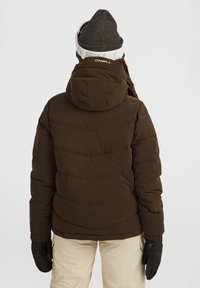 Bruine pufferjack met chevronsteek, aangehechte capuchon en elastische manchetten, gedragen met crèmekleurige broek en zwarte handschoenen.