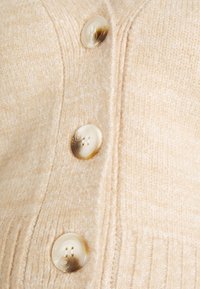 Cardigan bege em malha com tecido texturizado, bainha canelada proeminente e três grandes botões bege com padrões espiralados.
