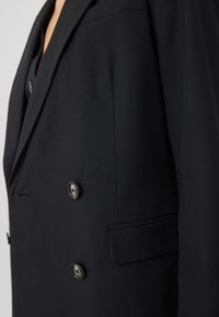 Černý dvouřadý blejzr s přiléhavým střihem. Obsahuje velké knoflíky a texturovanou látku. Viditelná boční kapsa.