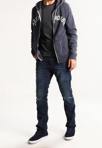 Man med marinblå hoodie med dragkedja som har texten "Abercrombie", mörkrandig t-shirt, slitna blå jeans, och marinblå sneakers, stående med händerna delvis i fickorna.