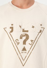 Felpa color crema in cotone con una grande grafica marrone a effetto usurato che presenta la parola "GUESS" e un design triangolare.