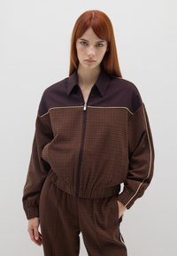 Bruine geruite zip-up jas met een donkere kraag en elastische tailleband. Heeft lange mouwen en een contrasterende crèmekleurige bies langs de schouders.