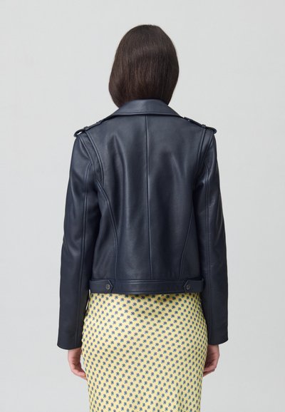Veste de moto en cuir noir avec épaulettes, coupe ajustée et boutons-pression à la taille. Portée par-dessus une jupe jaune à motifs.