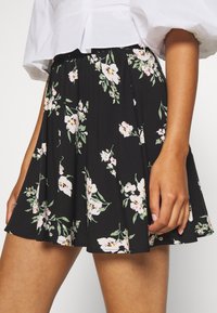 Jupe fleurie en tissu noir, avec un design plissé orné de fleurs blanches et roses. Taille élastique et longueur au-dessus du genou.