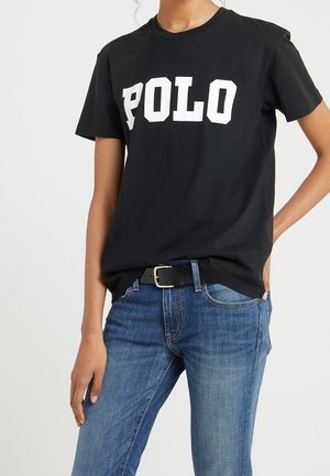 Persona que lleva una camiseta negra con texto blanco "POLO" y vaqueros azules, con una mano en el bolsillo y la otra a su lado.
