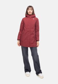 Cappotto isolato rosso con cappuccio, caratterizzato da un design essenziale e chiusura frontale con zip. Abbinato a jeans blu scuro e sneaker bianche.