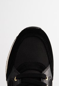 Black sneaker toe featuring mesh fabric, suede overlays, leather accents, and black laces with gold eyelets on a white background.