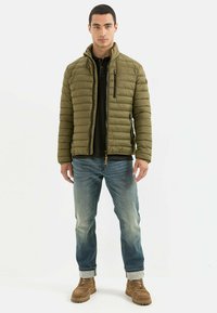 Grønne pufferjakke med horisontalt quiltet mønster, lynlåset front og sidelommer, båret over en sort skjorte, parret med blå jeans og brune støvler.