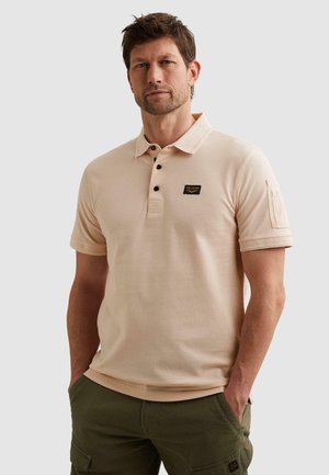 Mann mit kurzen braunen Haaren und Stoppelbart, trägt ein hellbeiges Polohemd mit schwarzen Knöpfen und olivgrünen Cargohosen, Hände in den Taschen.