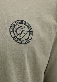 Μπεζ ύφασμα με στρογγυλό μαύρο λογότυπο που αναγράφει «Club Jack & Jones Since 1990» και περιβάλλει στυλιζαρισμένα επικαλυπτόμενα γράμματα CJ.