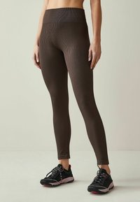 Bruine geribde leggings met een hoge tailleband, gemaakt van stretchstof; gecombineerd met zwarte sportschoenen met roze accenten en gestructureerde zolen.