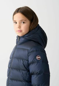 Námornícky modrá páperová bunda s kapucňou, s predným zipsom a nášivkou loga na ľavom ramene. Hladká textúra, prešívaný dizajn a priliehavý strih.