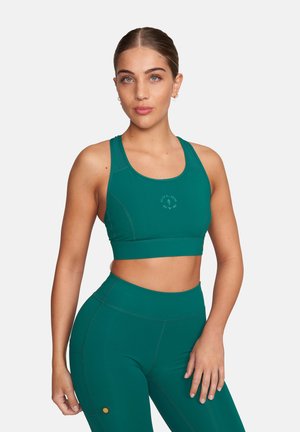 Kobieta w turkusowym sportowym biustonoszu i pasujących legginsach z wysokim stanem, stojąca z jedną ręką opartą na udzie, neutralne tło.