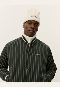 Black and white striped bomber jacket with a snap closure, ribbed collar, and embroidered logo. Cream knitted beanie completes the look.
