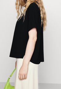 Svart, löst sittande topp med korta ärmar, kombinerad med en krämfärgad kjol. Personen håller en ljusgrön väska.