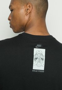 Sort bomulds T-shirt med et grafisk design på ryggen, der viser en tegneseriefigur, der holder en burger, og teksten "SOLE FOOD." Nike-logoet ovenfor.