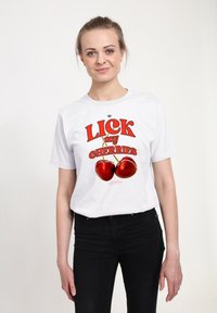 Žena s blond vlasy v ohonu, oblečená v bílém tričku s červeným nápisem "LICK my CHERRIES" a grafikou dvou třešní, v kombinaci s černými kalhotami.