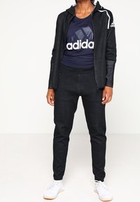 Chaqueta deportiva negra con cierre, detalles en blanco, camiseta de adidas azul marino, y pantalones oscuros. Zapatillas blancas completan el atuendo.