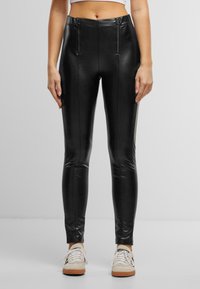 Leggings noirs en similicuir avec taille haute, présentant des accents de fermeture éclair à l'avant et des coutures verticales. Texture lisse, coupe ajustée, longueur cheville.