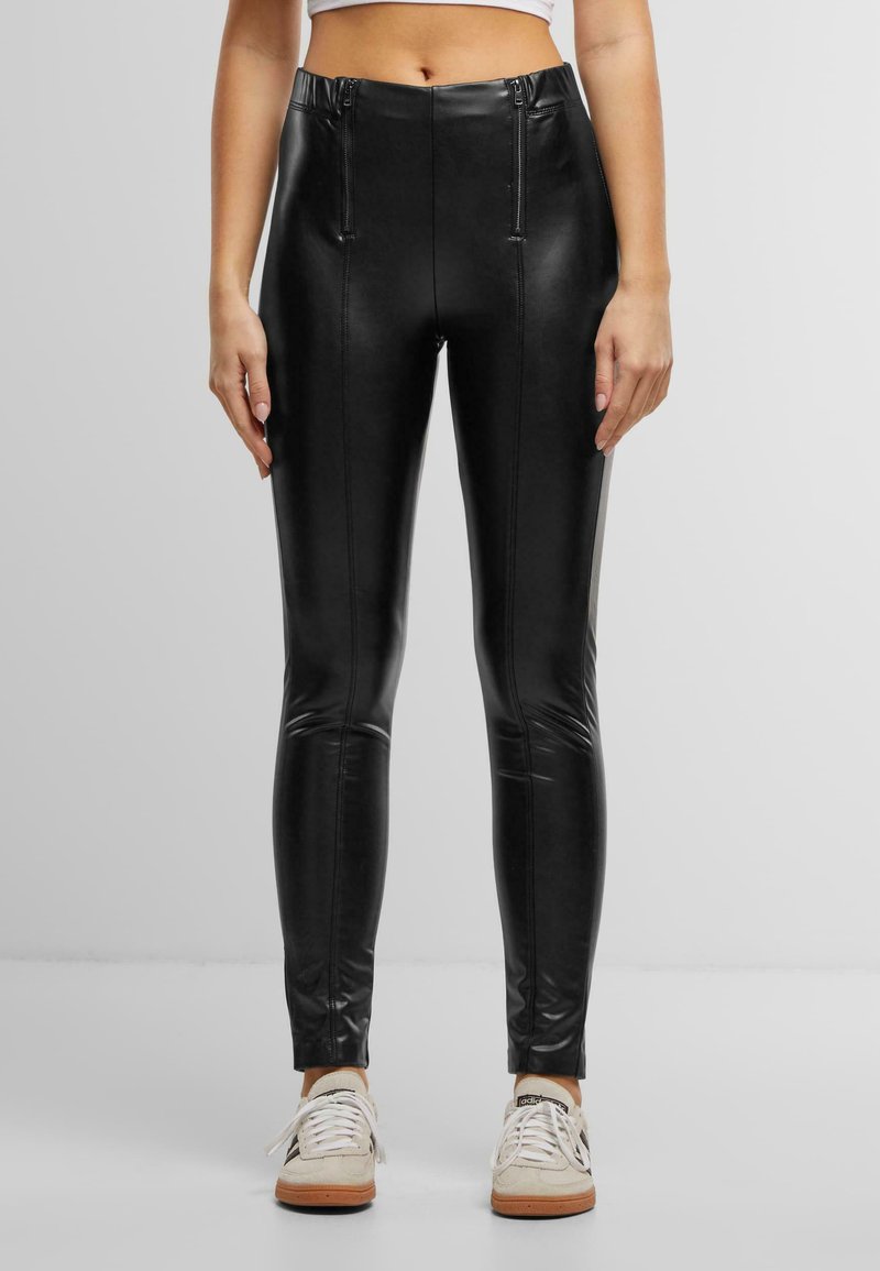 Leggings noirs en similicuir avec taille haute, présentant des accents de fermeture éclair à l'avant et des coutures verticales. Texture lisse, coupe ajustée, longueur cheville.