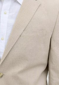Beige textured blazer with a slim fit. Features a single chest pocket and a two-button closure, paired with a white linen shirt underneath.