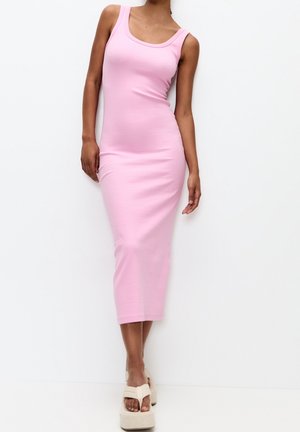 Femme portant une robe midi ajustée sans manches de couleur rose clair, associée à des sandales à plateforme beiges, debout devant un fond blanc.