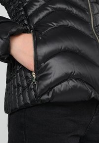 Main dans la poche zippée d'une doudoune matelassée noire portée sur un pantalon noir, mettant en valeur la texture de la veste et le détail de la fermeture éclair.