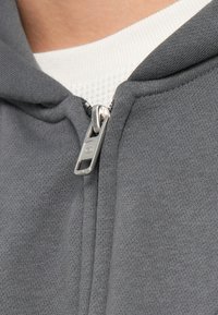Felpa grigia con zip, dal tessuto liscio, con zip argentata e colletto bianco sotto. Vista ravvicinata del tessuto.