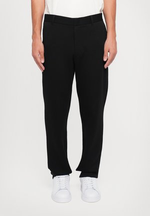 Black, loose-fitting trousers with a flat front, belt loops, and side pockets, paired with white sneakers. Textured fabric.