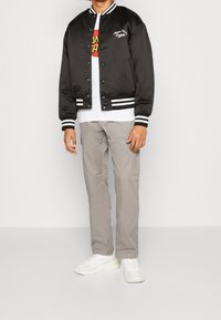 Zwarte satijnen bomberjack met witte gestreepte kraag en manchetten, voorzien van een logo op de borst, gecombineerd met beige cargobroek en witte sneakers.