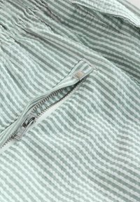Hellgrüner Stoff mit weißen Streifen, ausgestattet mit einem silbernen Reißverschluss und einem metallischen Zipper. Weiche Textur mit gerafften Details entlang der Kanten.