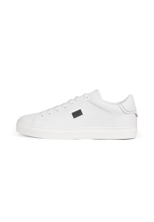 Zapatilla blanca de cuero con caña baja, cordones blancos, suela de goma y una pequeña placa metálica rectangular con el logo en el lateral.