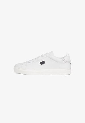 Zapatilla blanca de cuero con caña baja, cordones blancos, suela de goma y una pequeña placa metálica rectangular con el logo en el lateral.