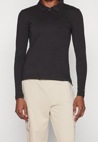 Person klädd i en svart långärmad polo med knappar och beige cargobyxor med sidofickor, stående mot en enfärgad bakgrund.