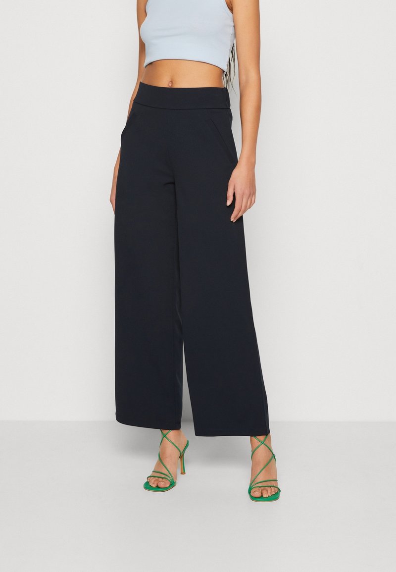 Femme portant un pantalon noir à jambes larges, un débardeur court bleu clair et des sandales à talons verts à brides, debout devant un fond uni.