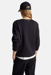 Schwarzer Sweatshirt mit Rundhalsausschnitt, langen Ärmeln und einer Nahtdetails auf der Rückseite. Getragen mit einer schwarzen Mütze und über einem weißen Hemd.