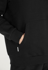 Käsi musta hupparin taskussa, jossa on ribbestähti hihansuissa ja vyötäröllä, näkyvät ompelu yksityiskohdat ja pieni valkoinen tunnus.