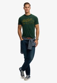 Camiseta de algodón verde con logotipo gráfico amarillo, vaqueros azul oscuro y una camisa a cuadros azul y blanca anudada a la cintura; zapatillas casuales.