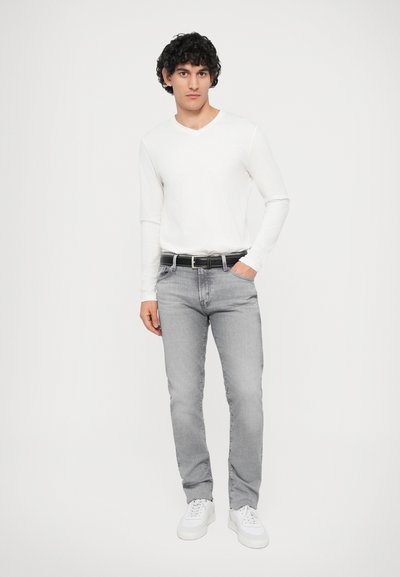 Graue Jeans aus Denim mit schmalem Schnitt, kombiniert mit einem weißen langärmeligen V-Ausschnitt-Shirt. Dazu ein schwarzer Gürtel und weiße Sneakers.