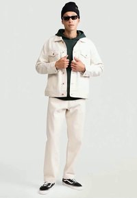 Mężczyzna poprawia jasnobeżową kurtkę na ciemnozielonym hoodie, ma na sobie beżowe spodnie, czarną czapkę beanie, okulary przeciwsłoneczne i czarno-białe sneakersy, stojąc.