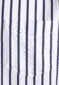 Weißer Stoff mit abwechselnd dunkelblauen vertikalen Streifen, mit einer quadratischen Tasche im unteren Bereich, einfarbige Textur und saubere Nähte.