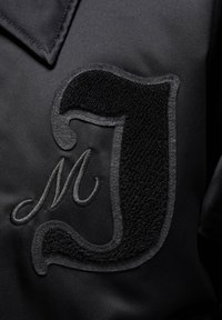 Czarna satynowa kurtka z teksturowanym czarnym aplikowanym literą "J" oraz haftowanym "M". Gładki materiał z kontrastowymi detalami na powierzchni.