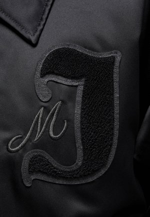 Veste en satin noir avec un lettrage en appliqué texturé noir en forme de "J" et un "M" brodé. Tissu lisse avec des détails de surface contrastés.