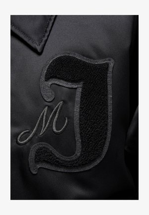 Sorte satinjakke med en struktureret sort applikation af bogstavet "J" og broderet "M". Glat stof med kontrasterende overfladedetaljer.