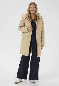 Manteau matelassé beige avec capuche, doté de deux poches avant et d'une fermeture éclair. Associé à un jean large foncé et des sneakers blanches.