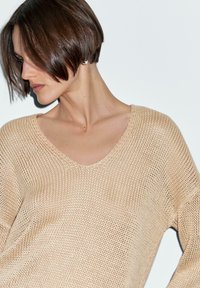 Beżowy sweter robiony na drutach z szerokim dekoltem w kształcie litery V, luźnym kroju i teksturowanym wzorem. Materiał wydaje się lekki i przewiewny.