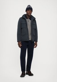 Chaqueta de abrigo azul marino con múltiples bolsillos frontales, camiseta de rayas, pantalones oscuros y botas negras con cordones, rematada con un gorro de lana azul marino.