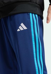 Primer plano de pantalones deportivos azules con tres rayas azul claro y logo blanco en el muslo, usados con camiseta negra, mano visible al lado.