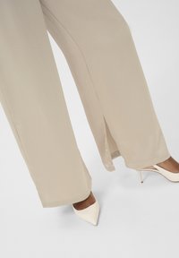 Pantalons beige à jambes larges avec une texture côtelée subtile, présentant des fentes à l'ourlet. Associés à des talons blancs à bout pointu.